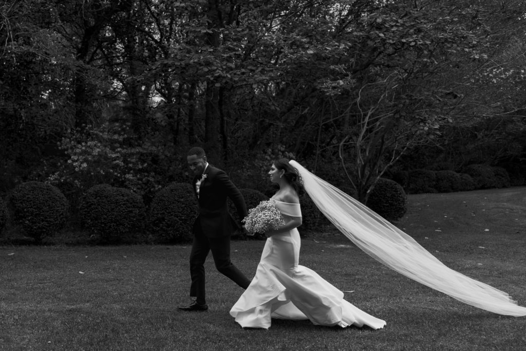 Wedding couple runs through grassy lawn surrounded by large trees