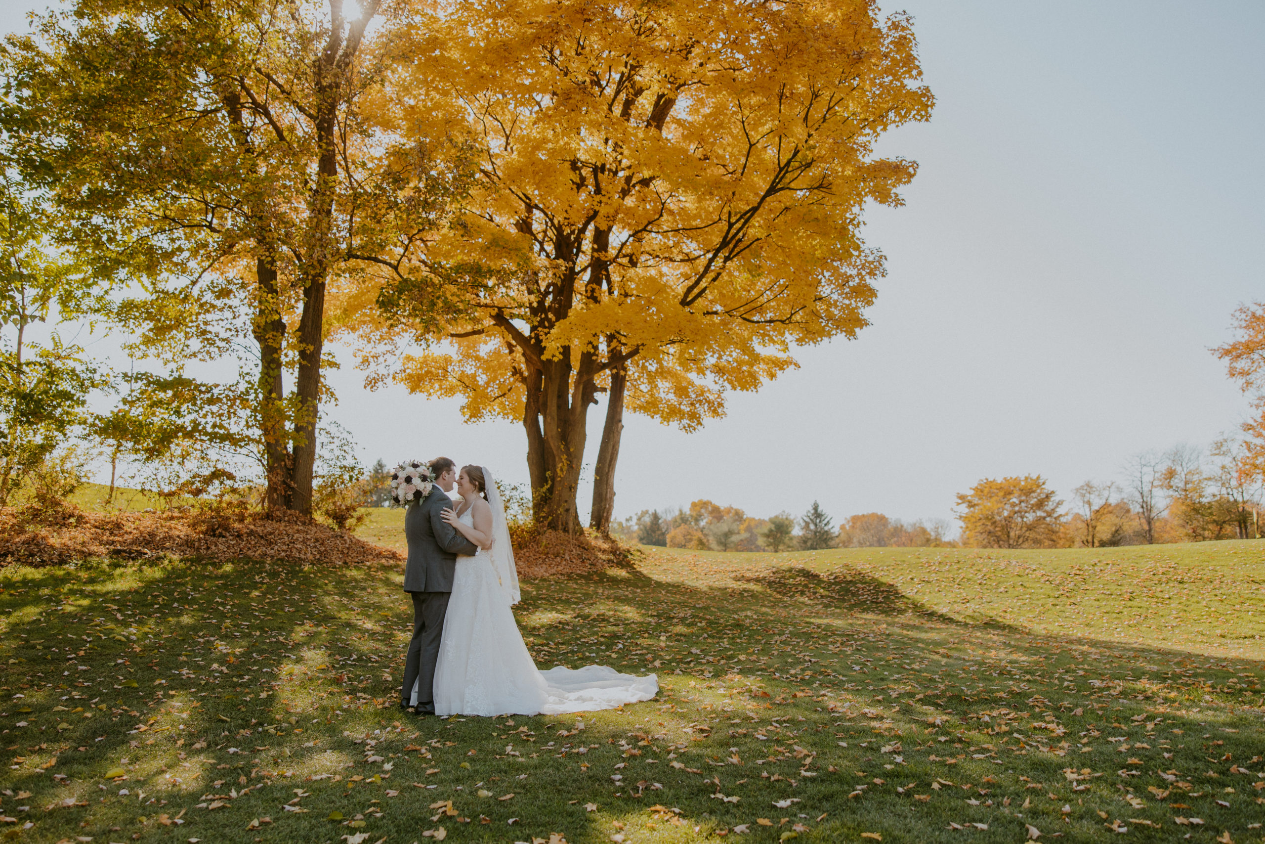 Couple stands below gorgeous tree with fall leaves.